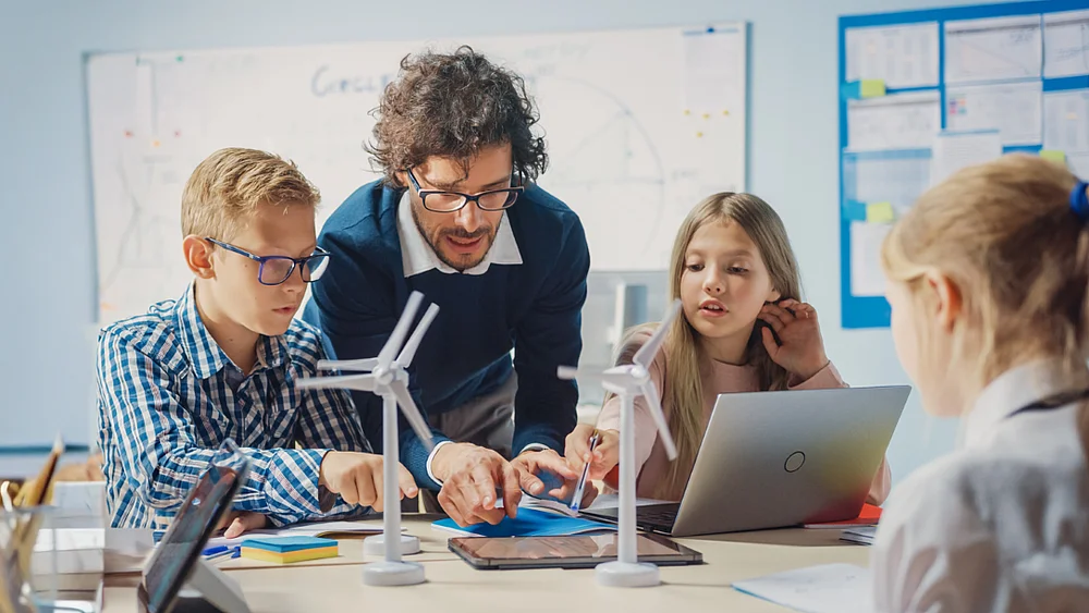 Kinder arbeiten gemeinsam mit Lehrkraft an einem Energiewende-Projekt mit Windkraftmodellen und digitalen Lernmedien.