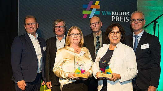 Platz 2: Forum Anja Niedringhaus Höxter Gruppenfoto vor einer Fotowand von Vereinsmitgliedern des Forum Anja Niedringhaus mit dem 2. Hauptpreis des WWKulturpreis26.