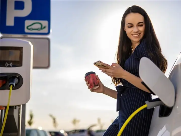 Eine Person lädt ein Elektroauto an einer Wallbox und hält dabei ein Smartphone und einen Kaffeebecher.