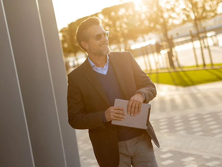 Westfalen Weser Fernwärme Herford Ein Geschäftsmann steht an einem Gebäude und hält Unterlagen in den Händen. Er lächelt und die Sonne scheint im Hintergrund.