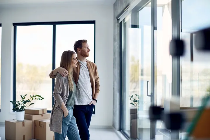 Ein Paar steht in der neuen Wohnung an der Terrassentür und schaut raus. Die beiden scheinen sich im Umzug zu befinden, da man im Hintergrund Kartons sieht.