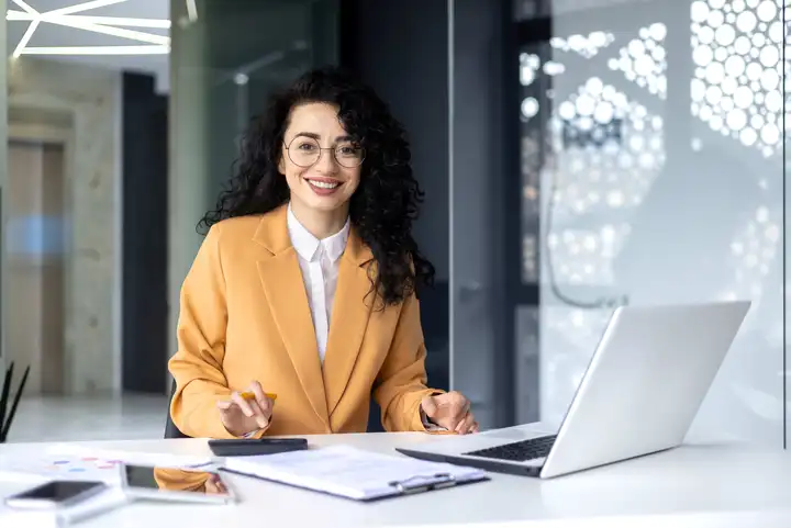 Eine Frau sitzt an einem Schreibtisch und arbeitet an ihrem Laptop und lächelt in die Kamera.