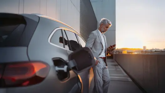 Eine Person im Anzug lädt ein Elektroauto und nutzt ein Smartphone auf einem Parkplatz bei Sonnenuntergang.