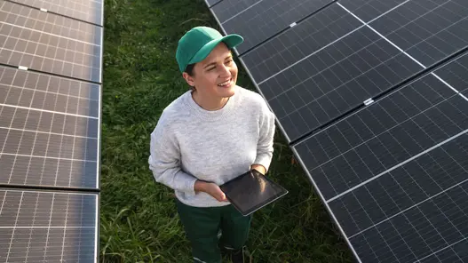 Eine Frau läuft durch ein Feld mit Photovoltaikanlagen mit einem Tablet in der Hand und lächelt.