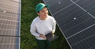 Eine Frau läuft durch ein Feld mit Photovoltaikanlagen mit einem Tablet in der Hand und lächelt.