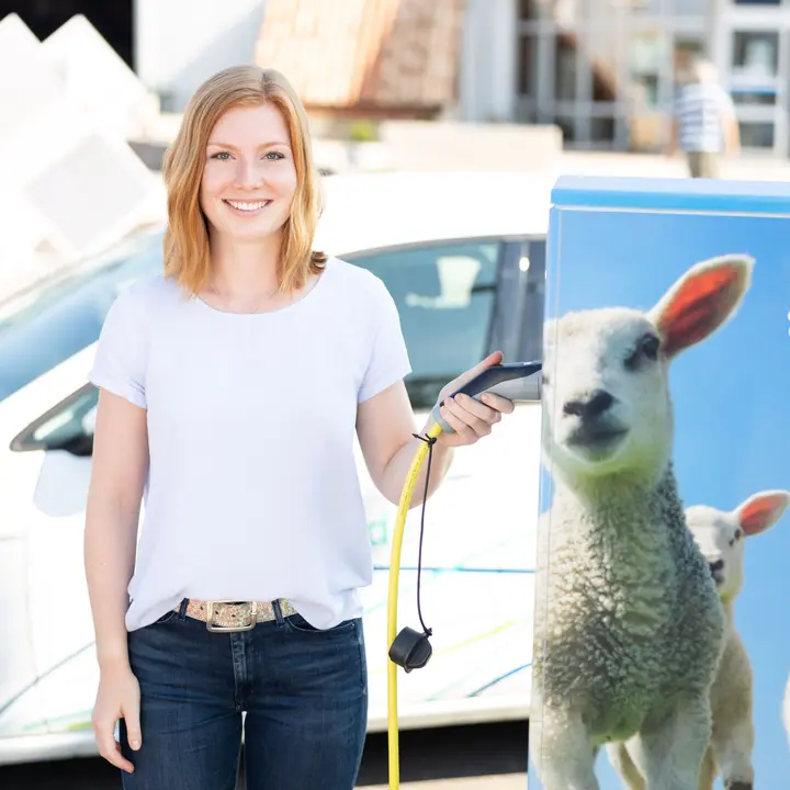 Eine Person verbindet ein Elektroauto mit einer Ladesäule, die mit einem Schafmotiv und E-Auto-Symbol gestaltet ist.