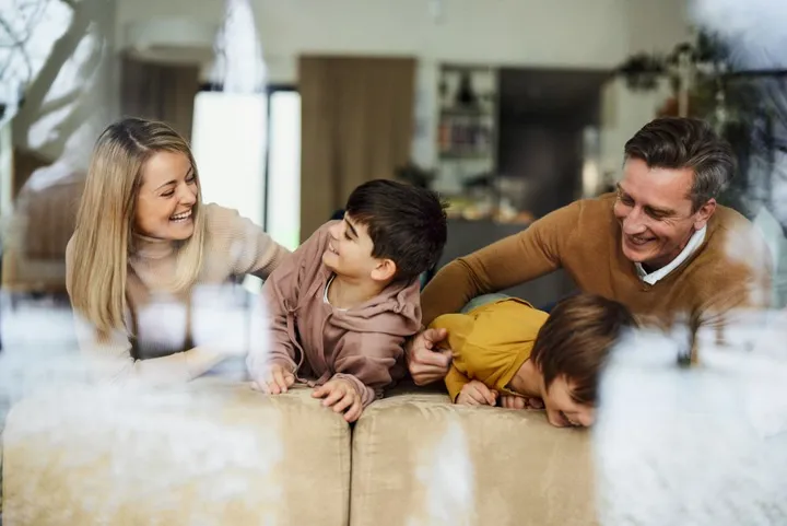 Eine vierköpfige Familie lehnt auf der Lehne der Couch und spielt miteinander. Der Vater kitzelt die Tochter. Alle lachen.