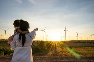 Eine Frau mit einem Kind auf dem Arm steht auf einer Wiese und zeigt auf einen Windpark in der Ferne.