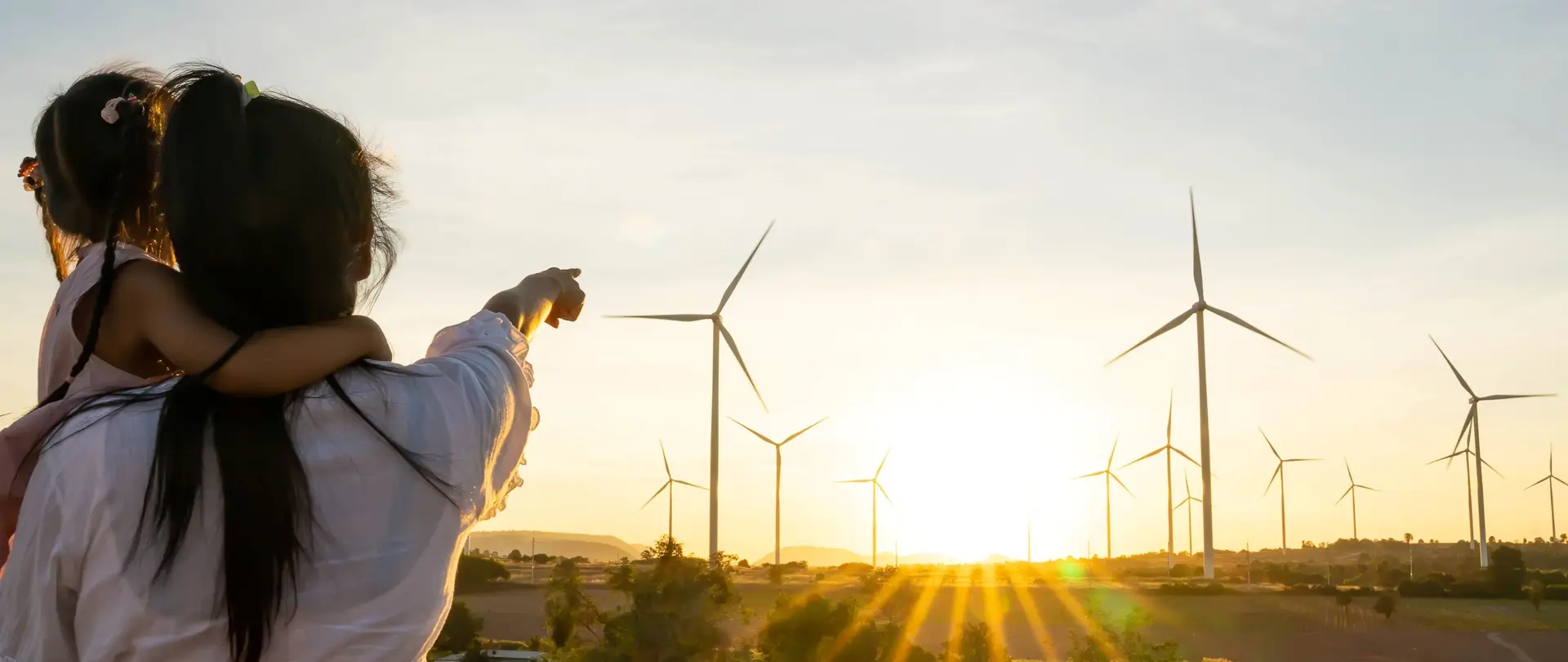 Eine Frau mit einem Kind auf dem Arm steht auf einer Wiese und zeigt auf einen Windpark in der Ferne.