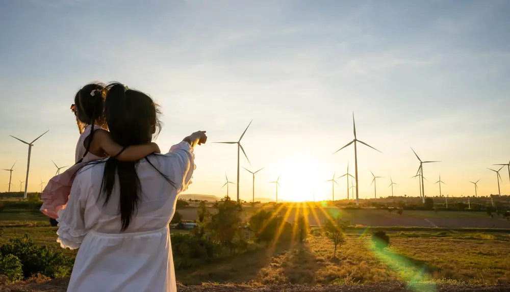 Eine Frau mit einem Kind auf dem Arm steht auf einer Wiese und zeigt auf einen Windpark in der Ferne.