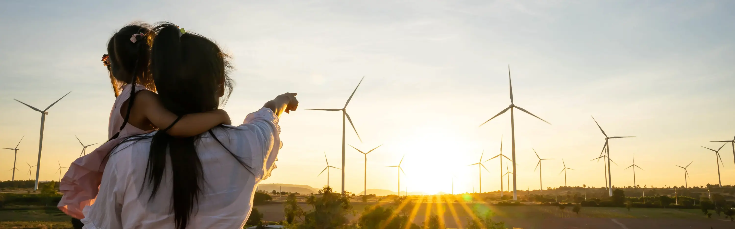 Eine Frau mit einem Kind auf dem Arm steht auf einer Wiese und zeigt auf einen Windpark in der Ferne.