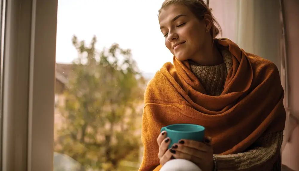 Eine in der Decke eingewickelte Frau sitzt auf einer Fensterbank, hält eine Tasse Kaffee in den Händen und genießt den Moment. Ihre Augen sind geschlossen.