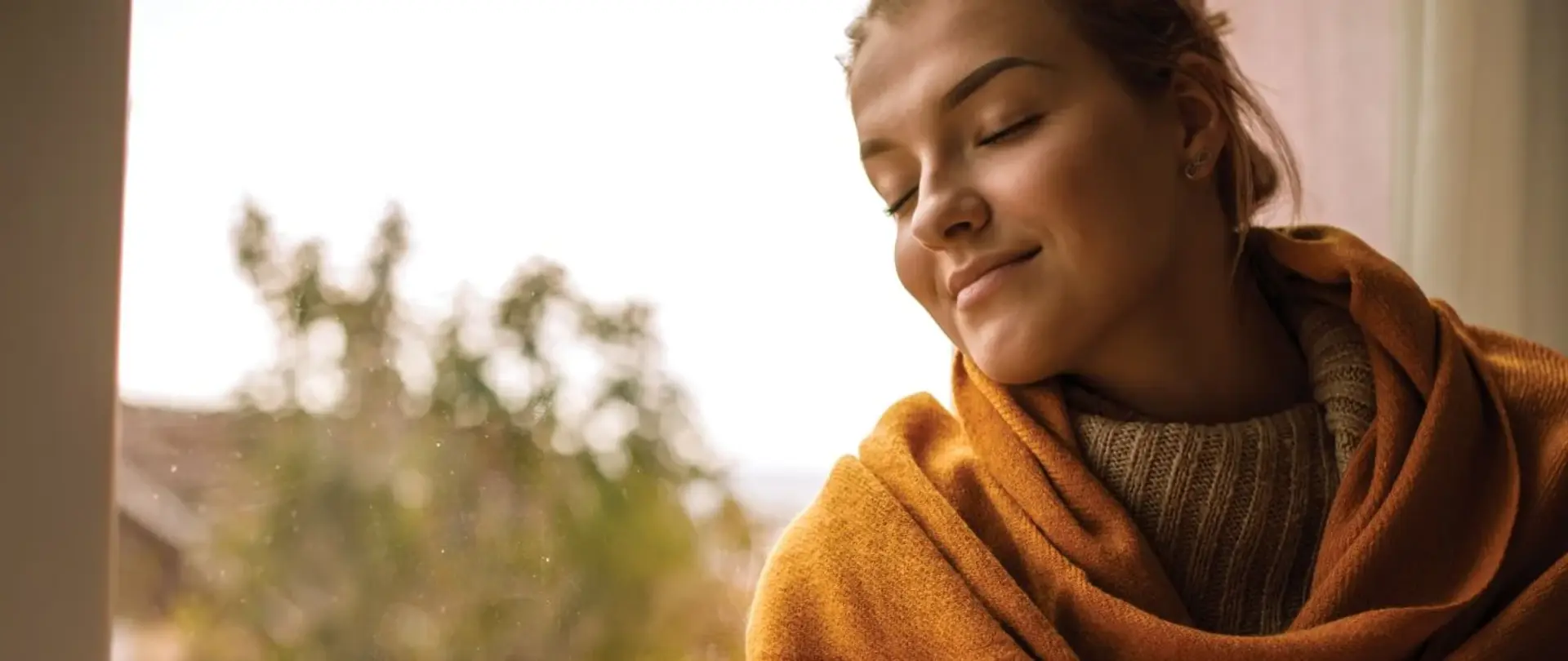 Eine in der Decke eingewickelte Frau sitzt auf einer Fensterbank, hält eine Tasse Kaffee in den Händen und genießt den Moment. Ihre Augen sind geschlossen.