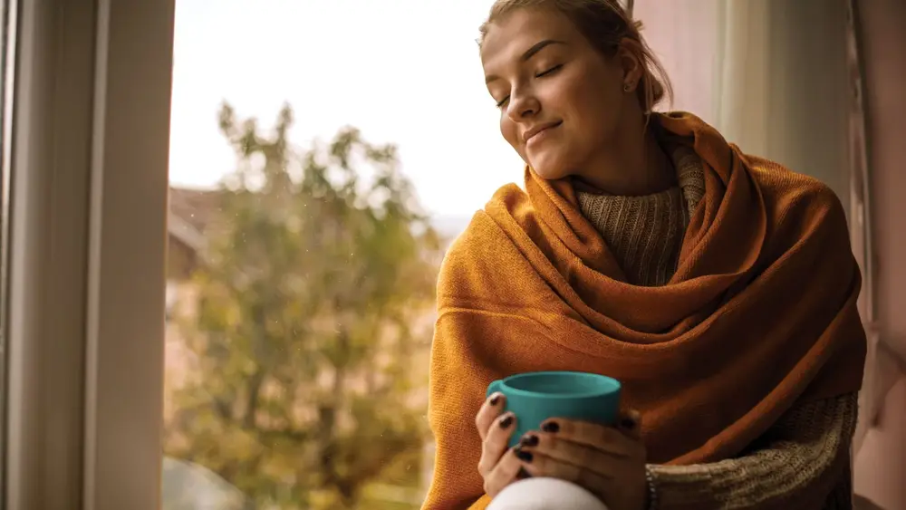 Eine in der Decke eingewickelte Frau sitzt auf einer Fensterbank, hält eine Tasse Kaffee in den Händen und genießt den Moment. Ihre Augen sind geschlossen.