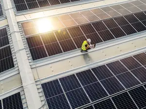 Eine Person mit Helm und Warnweste arbeitet auf einem Solardach mit Laptop zwischen reflektierenden Solarpanels.