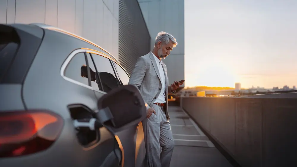 Eine Person im Anzug lädt ein Elektroauto und nutzt ein Smartphone auf einem Parkplatz bei Sonnenuntergang.