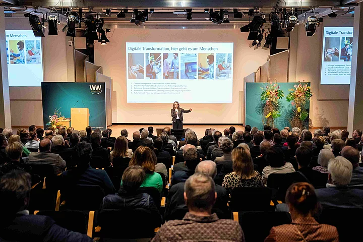 Westfalen Weser Forum 2025 - Publikum und Referentin Man sieht das Publikum von hinten mit Fokus auf die Referentin Anna Kopp, die auf der Bühne steht und einen Vortrag hält.