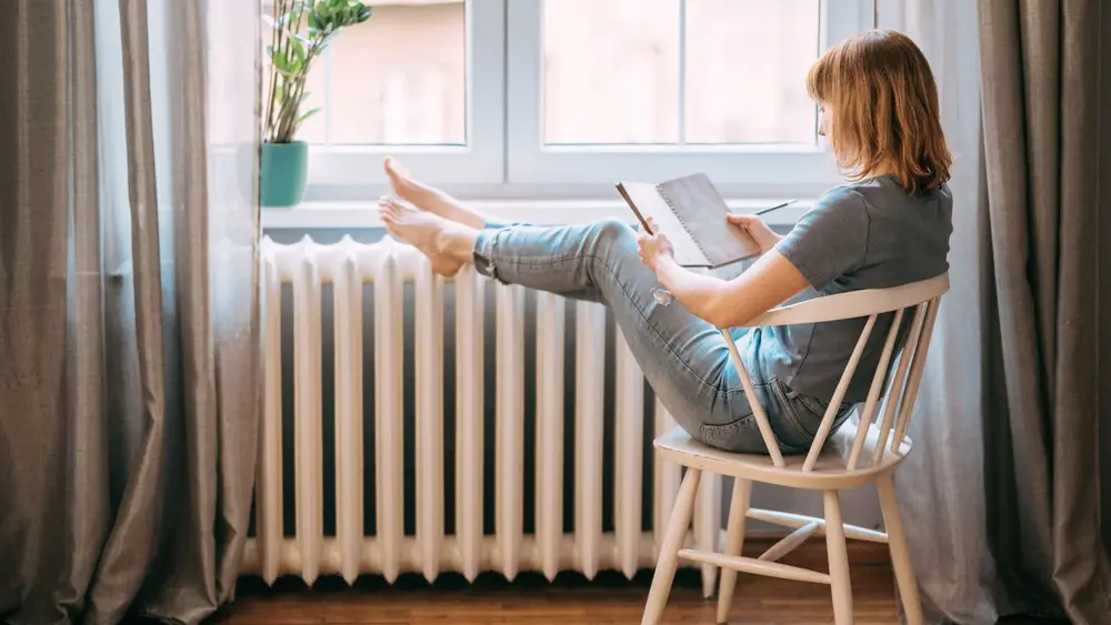 Eine Frau sitzt auf einem Holzstuhl und liest ein Buch. Ihre Füße wärmt sie auf der Heizung. Auf der Fensterbank steht eine Pflanze
