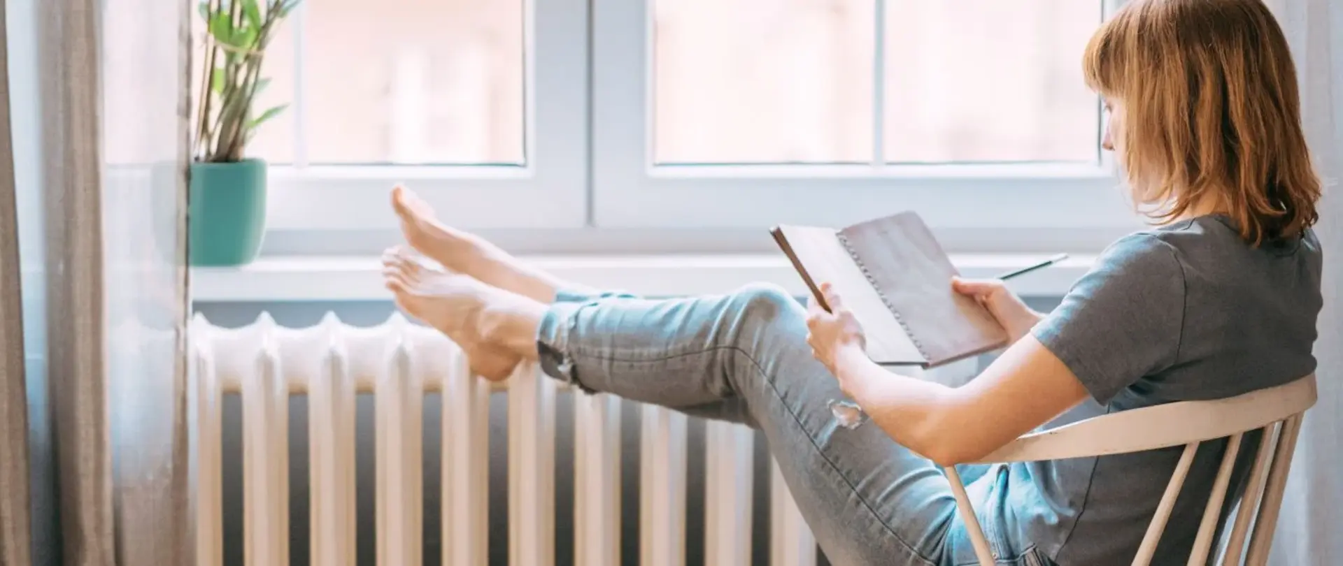 Eine Frau sitzt auf einem Holzstuhl und liest ein Buch. Ihre Füße wärmt sie auf der Heizung. Auf der Fensterbank steht eine Pflanze