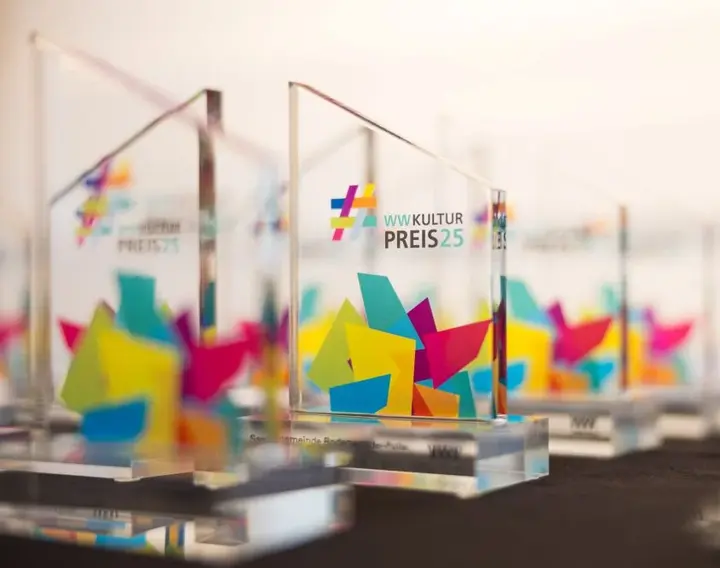 Mehrere Preise des WWKulturPreises stehen auf einem schwarzen Tisch. Die Preise sind aus Plexiglas und mit dem WWKulturpreis Logo versehen 