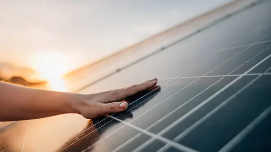 Eine Hand liegt auf einem PV-Modul und im Hintergrund geht die Sonne langsam unter.