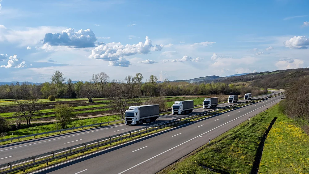 Mehrere LKW fahren hintereinander über eine Autobahn in ländlicher Umgebung.