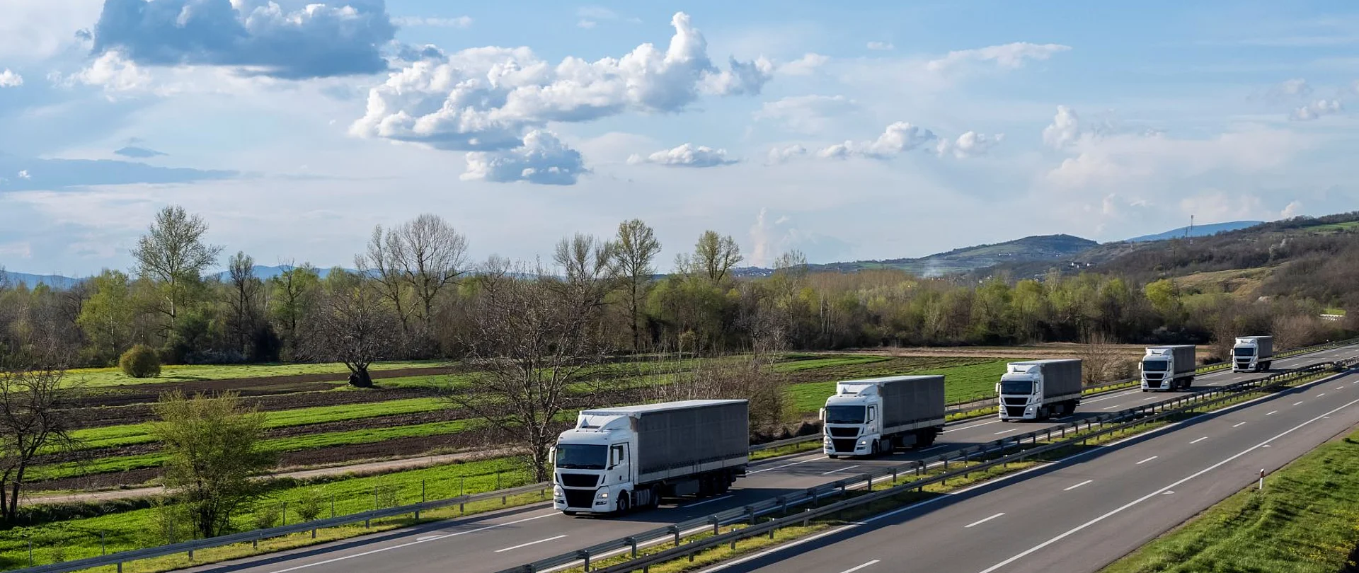Mehrere LKW fahren hintereinander über eine Autobahn in ländlicher Umgebung.