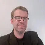 Ein freundlich lächelnder Mann im schwarzen Jackett und mit Brille steht vor einer weißen Wand.