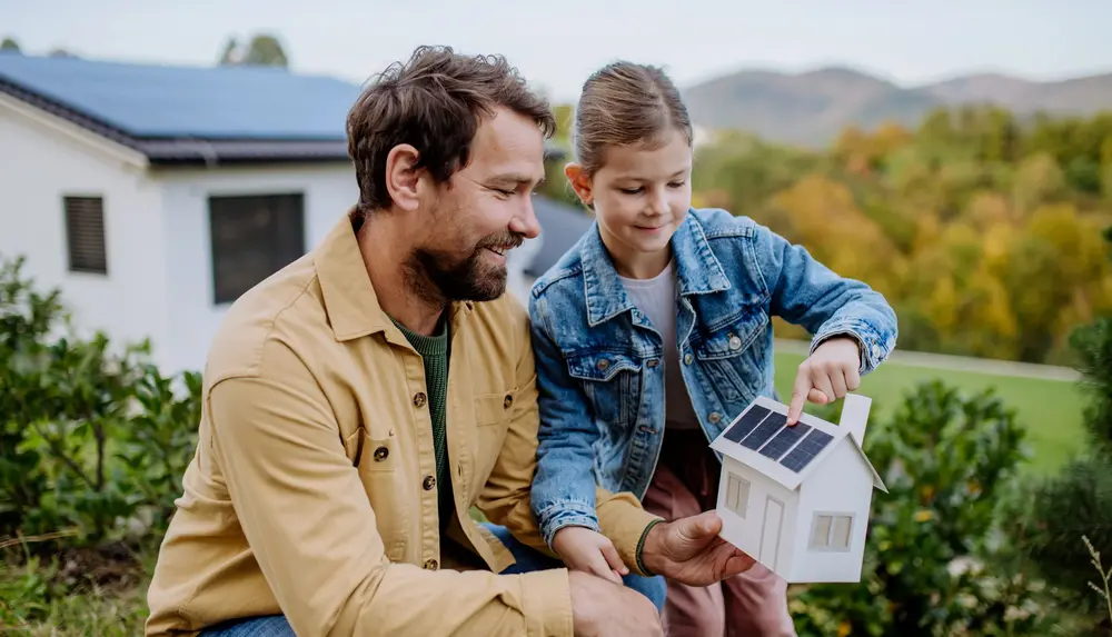 Eine erwachsene Person und ein Kind betrachten gemeinsam ein Modellhaus mit Solarpanelen vor einem modernen Haus mit Solardach.