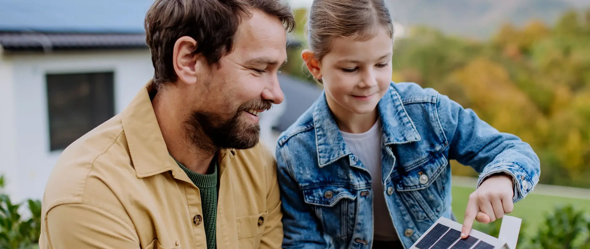 Eine erwachsene Person und ein Kind betrachten gemeinsam ein Modellhaus mit Solarpanelen vor einem modernen Haus mit Solardach.