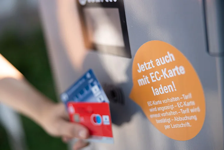 ine Person hält zwei Girokarten an dieLadesäule für ein Elektroauto.