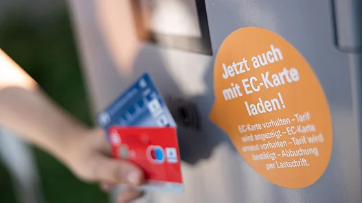 ine Person hält zwei Girokarten an dieLadesäule für ein Elektroauto.