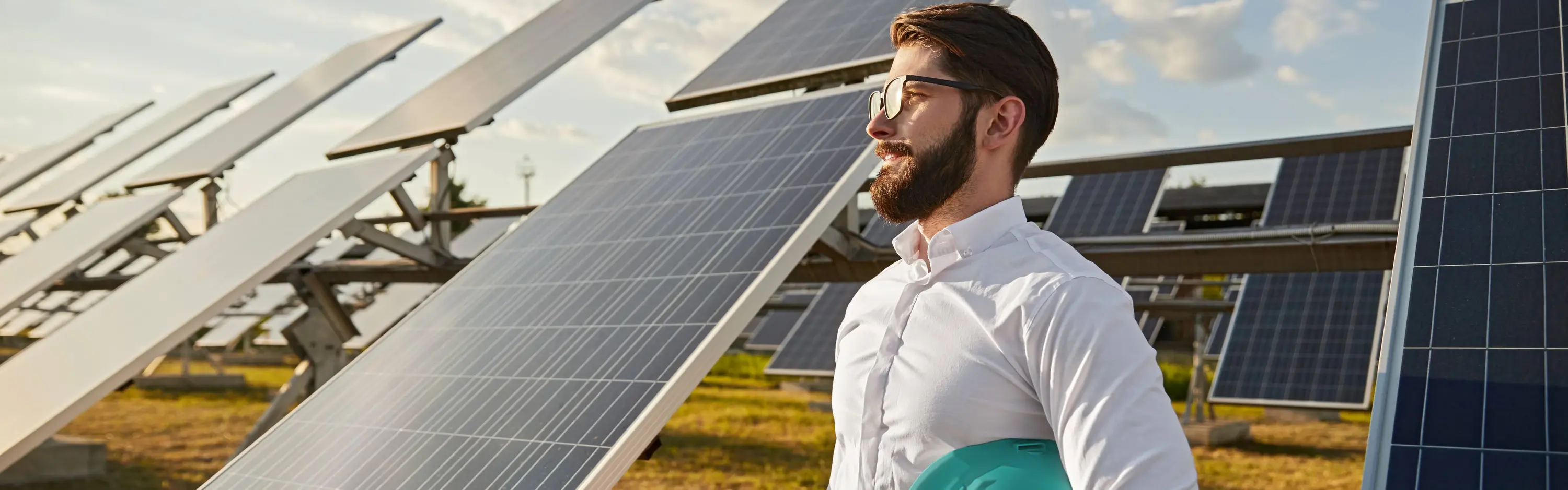 Ein Mann in Business-Kleidung und einem Helm unter dem Arm steht auf einem Feld vor PV-Freiflächenanlagen und lächelt in die Ferne.