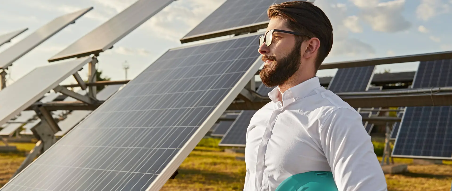 Ein Mann in Business-Kleidung und einem Helm unter dem Arm steht auf einem Feld vor PV-Freiflächenanlagen und lächelt in die Ferne.
