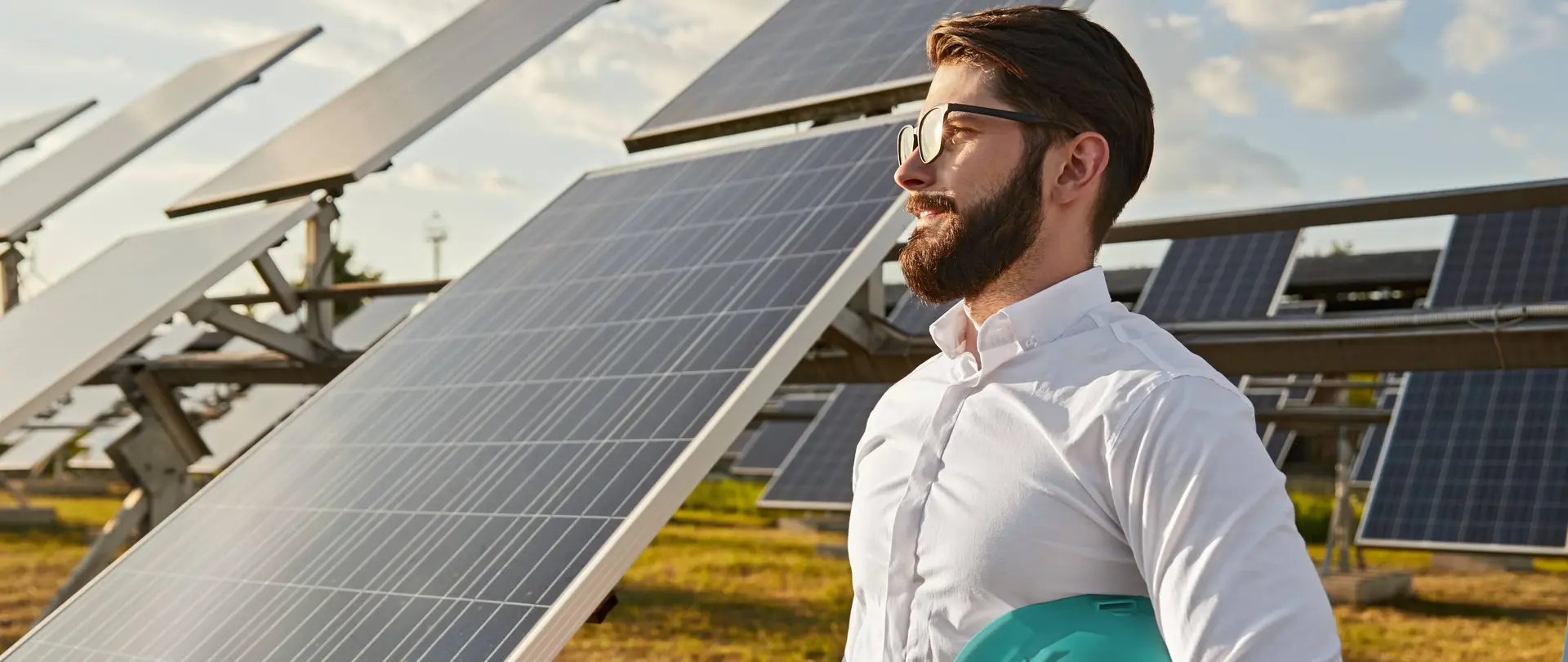 Ein Mann in Business-Kleidung und einem Helm unter dem Arm steht auf einem Feld vor PV-Freiflächenanlagen und lächelt in die Ferne.