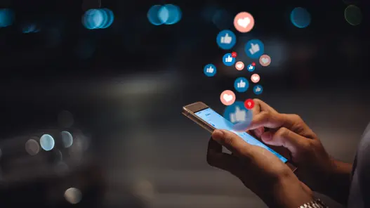 Eine Person hält ein Smartphone, auf dem Social-Media-Symbole wie Daumen hoch und Herzen erscheinen.
