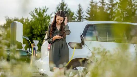 Eine Person lädt ein weißes Elektroauto an einer Wallbox und nutzt dabei ein Smartphone zur Steuerung.