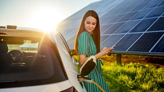 Eine Person steht mit Smartphone neben einem Elektroauto, das bei Sonnenuntergang mit Solarstrom geladen wird.