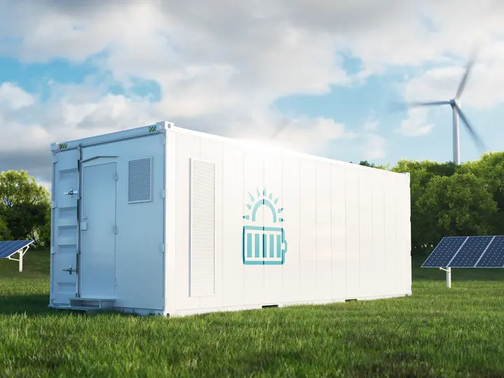 Ein weißer Batteriegroßspeicher mit steht vor Solarpaneelen und einem Windrad auf einer Wiese.