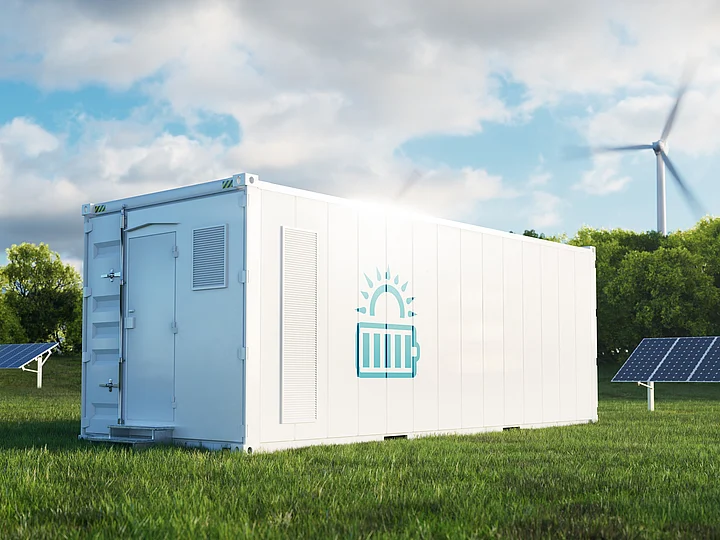 Ein weißer Batteriegroßspeicher mit steht vor Solarpaneelen und einem Windrad auf einer Wiese.