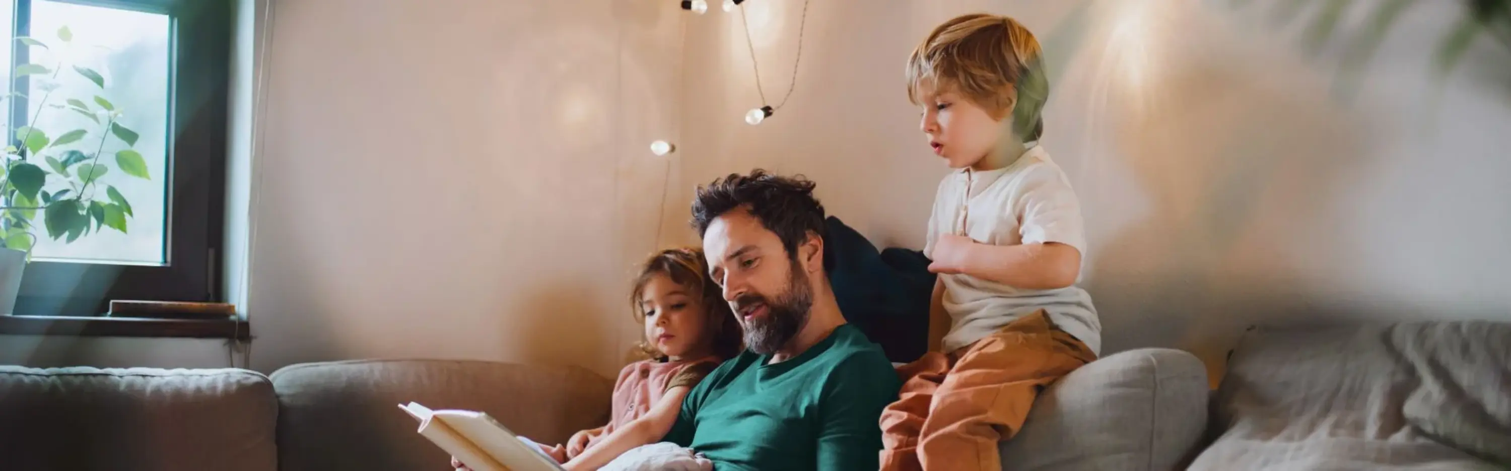 Ein Vater sitzt mit seinen beiden Kindern auf dem Sofa und liest aus einem Buch vor. Die Kinder hören gespannt zu und schauen mit in das Buch