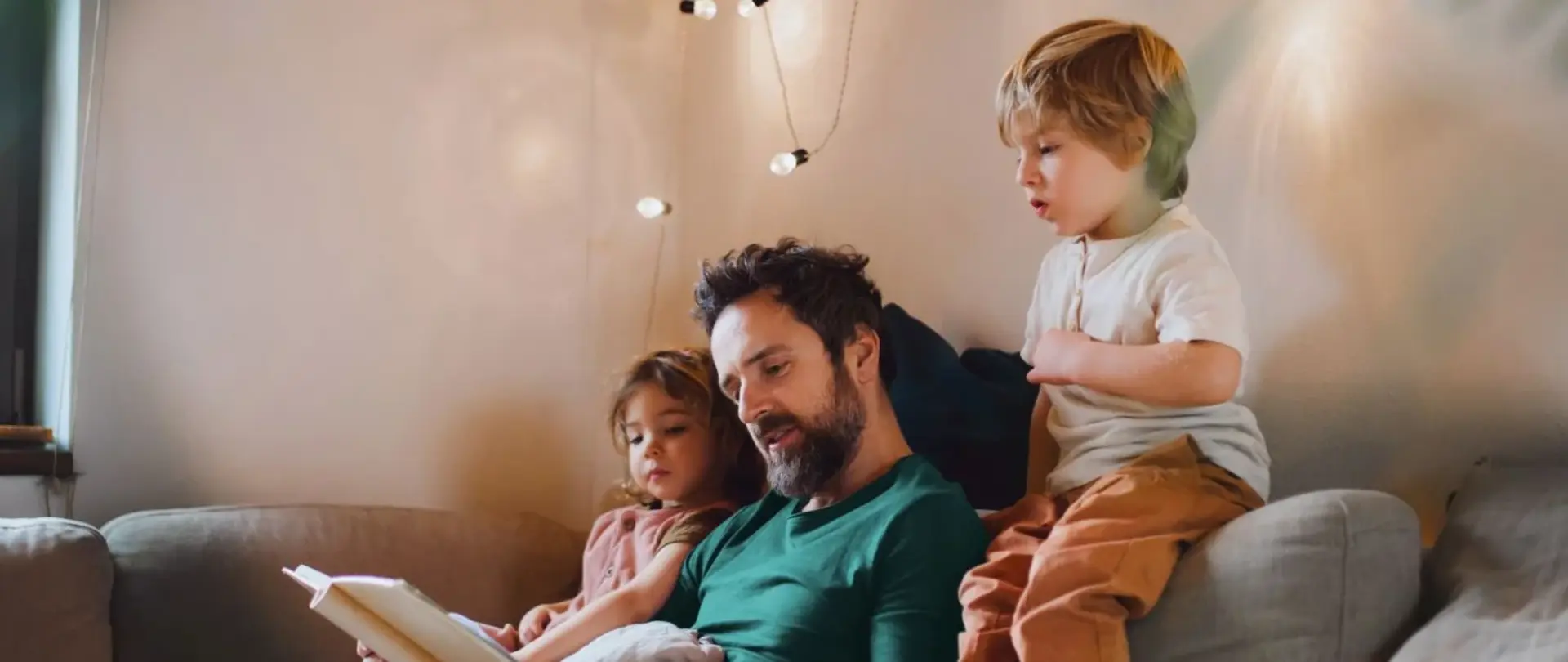 Ein Vater sitzt mit seinen beiden Kindern auf dem Sofa und liest aus einem Buch vor. Die Kinder hören gespannt zu und schauen mit in das Buch
