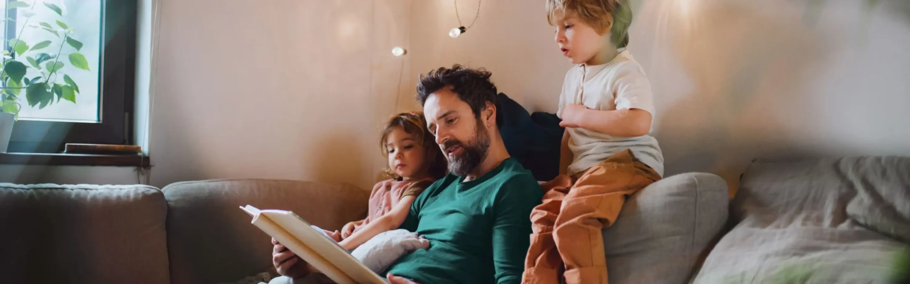 Ein Vater sitzt mit seinen beiden Kindern auf dem Sofa und liest aus einem Buch vor. Die Kinder hören gespannt zu und schauen mit in das Buch