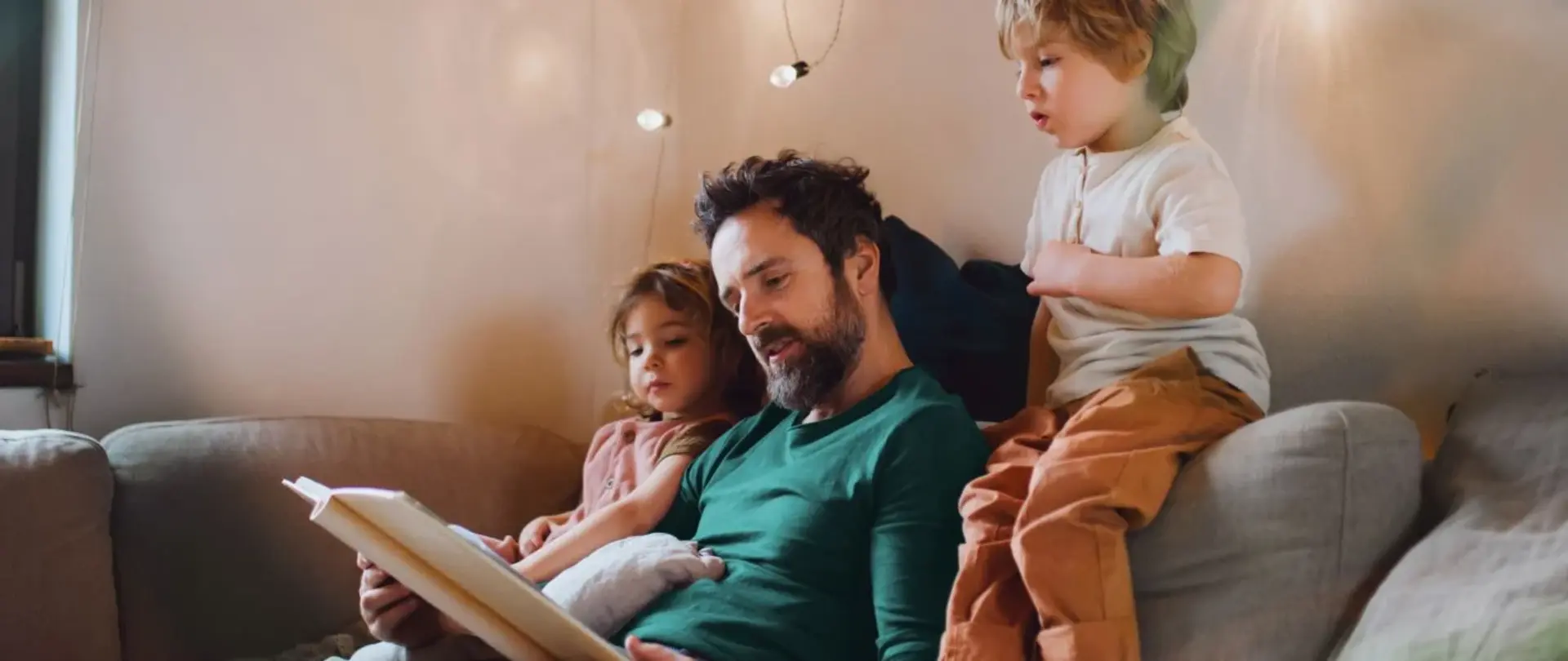 Ein Vater sitzt mit seinen beiden Kindern auf dem Sofa und liest aus einem Buch vor. Die Kinder hören gespannt zu und schauen mit in das Buch