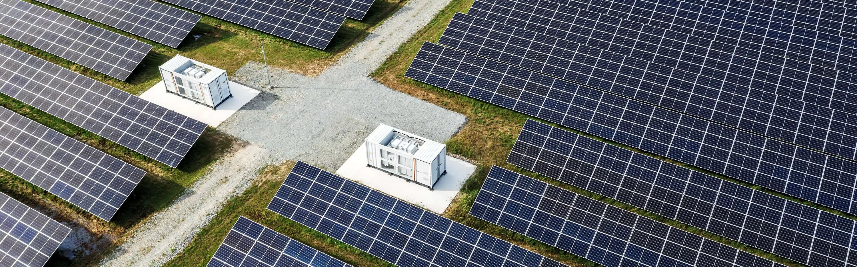 Eine große Solaranlage mit Batteriespeichern und Schotterweg verläuft durch grüne Flächen mit Solarpaneelen.