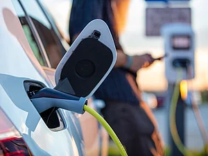 Elektroauto wird an einer öffentlichen Ladestation geladen. Ein angeschlossenes Ladekabel verbindet das Fahrzeug mit der Ladesäule. EIne Frau bedient im Hintergrund Ihr Smartphone.