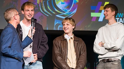 Die Band Motion hält eine Urkunde für den Start-Bonus des WWKulturSTARTER26 in der Hand, während der Moderator Ingo Börchers ein Interview auf der Bühne führt.