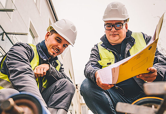 Bild mit zwei Personen kniend auf einer Baustelle in Arbeitskleidung mit Warnweste 
