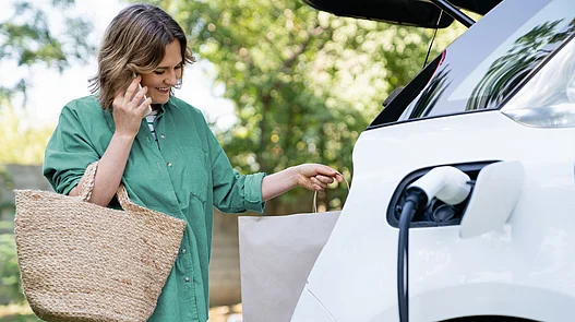Eine Person mit Shoppingtasche greift zum Ladestecker eines Elektroautos und nutzt gleichzeitig ein Smartphone.