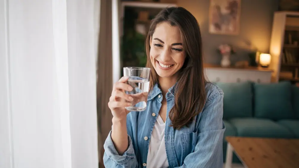 Eine Frau steht am Fenster und hält ein Glas Wasser in der Hand. Sie trägt ein Jeans-Hemd und eine weiße Bluse